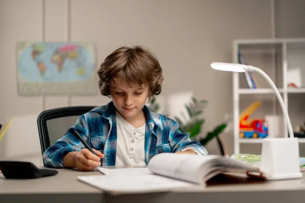 Çocuk odasında beyaz kazaklı ve mavi kareli tişörtlü bir okul çocuğu ders çalışmalarını not defterine yazarak yapıyor.
