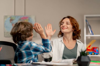 Çocuklar odasında beyaz kazaklı ve mavi kareli tişörtlü bir okul çocuğu annesiyle ödev yapıyor. Her şey birbirine beşlik çakmaktan ibaret.