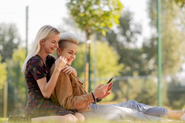 Yazın çimen kareli battaniyenin üzerinde oturmuş birbirine aşık bir çiftin sosyal ağlarda rahatlamasını seyreden güzel bir lezbiyen çift.