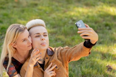 Yazın çimen kareli battaniyenin üzerinde oturup birbirine aşık bir çiftin telefonda rahatlarken fotoğraflarını çeken güzel lezbiyen bir çift.