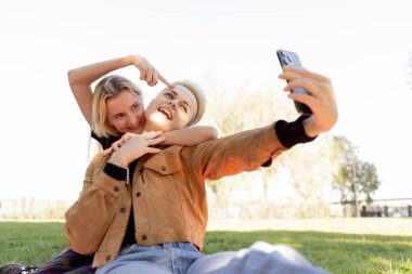 Yazın çimen kareli battaniyenin üzerinde oturup birbirine aşık bir çiftin telefonda rahatlarken fotoğraflarını çeken güzel lezbiyen bir çift.