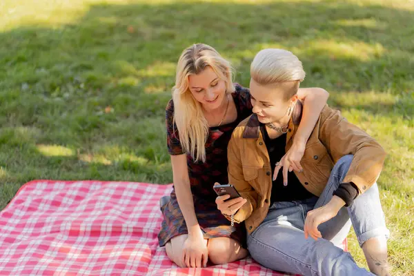 Yazın çimen kareli battaniyenin üzerinde oturmuş birbirine aşık bir çiftin sosyal ağlarda rahatlamasını seyreden güzel bir lezbiyen çift.