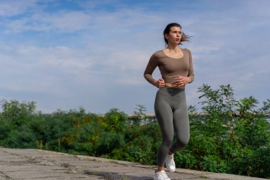 Şehir merkezindeki nehir kıyısındaki sokakta beton bir yolda gri spor takım elbiseli sağlıklı bir kız sabah koşusu yapıyor.