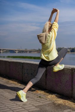 Şehir merkezindeki nehir kıyısındaki sokakta beton bir yolda sarı spor takım elbiseli, sağlıklı yaşam tarzı ısınma figürlü, mükemmel olmayan bir kız.