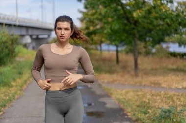 Caddeyi kapatın şehir merkezindeki nehir kıyısında beton bir yolda gri spor takım elbiseli, sağlıklı bir kız sabah koşusu yapıyor.