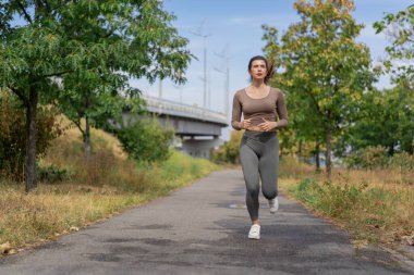 Şehir merkezindeki nehir kıyısındaki sokakta beton bir yolda gri spor takım elbiseli sağlıklı bir kız sabah koşusu yapıyor.