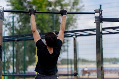 Parkta, yatay barların yanında siyah şortlu esmer ve siyah tişörtlü hoş bir adam yatay barda barfiks çekiyor sağlıklı yaşam tarzı kas pompalıyor.