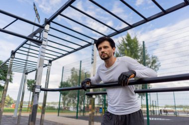 Yatay barların yanındaki parkta siyah şortlu ve beyaz tişörtlü yakışıklı bir esmer sağlıklı bir yaşam tarzının ardından yatay barın yanında dinleniyor.