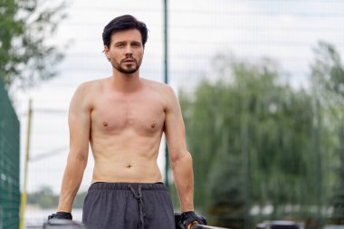 Yatay barların yanındaki parkta siyah şortlu yakışıklı bir esmer ve yatay barda şınav çeken ve kaslarını şişiren tişörtsüz bir adam.