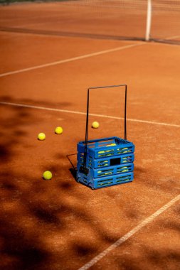 Profesyonel bir tenis kortunda kırmızı kilden bir yüzey çizilmiş bölgelerle tenis topları ve taşaklı bir sepet vardır.