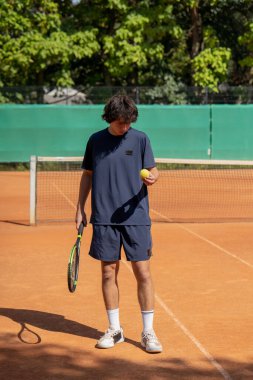 Profesyonel tenis kortunda genç kıvırcık adam elinde tenis topuyla ayakta duruyor.