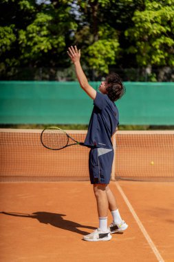 Profesyonel tenis kortunda genç kıvırcık çocuk teknik olarak oyunun ilk servisini yapıyor.