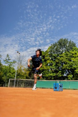 Profesyonel tenis kortunda genç kıvırcık çocuk teknik olarak oyunun ilk servisini yapıyor.