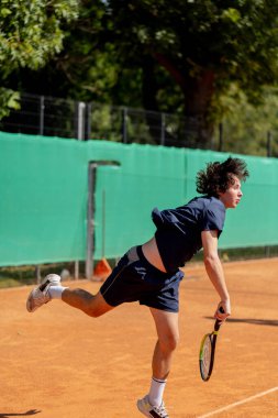 Profesyonel tenis kortunda genç kıvırcık çocuk teknik olarak oyunun ilk servisini yapıyor.