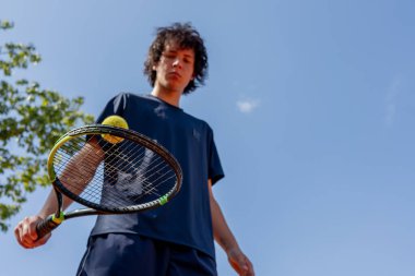 Profesyonel tenis kortuna yakın çekim. Genç kıvırcık çocuk tenis raketiyle topa vuruyor. Maç için profesyonel hazırlık.