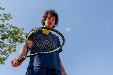 Profesyonel tenis kortuna yakın çekim. Genç kıvırcık çocuk tenis raketiyle topa vuruyor. Maç için profesyonel hazırlık.