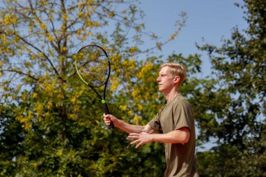 Profesyonel tenis kortunda genç sarışın bir adam tenis topuna vuruyor.