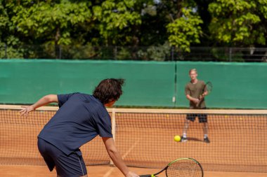 Profesyonel tenis kortunda genç kıvırcık çocuk sarışın rakip çiftle tenis oynuyor aktif dinlenme antrenmanı yapıyor raketle servis yapıyor.