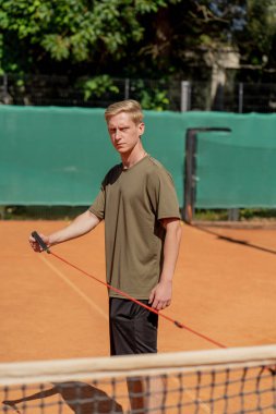 Profesyonel bir tenis kortunda kalenin yanında genç bir sarışın antrenör özel elastik bir bantla kollarının kaslarını güçlendirmek için antrenman yapıyor.
