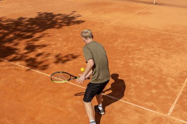 Profesyonel tenis kortunda genç sarışın bir adam tenis topuna vuruyor.