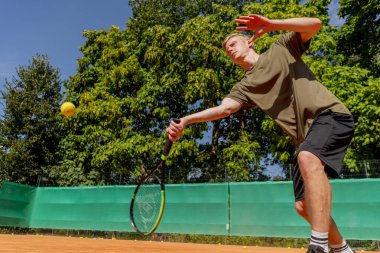 Profesyonel tenis kortunda genç sarışın bir adam tenis topuna vuruyor.