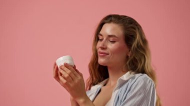 Stüdyonun güzellik çekimini kapat pembe arka planda uzun saçlı kız iç çamaşırı ve mavi gömlekli doğal güzellik kavanozuyla poz ver.