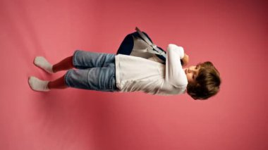 vertical video in a photo studio on a pink background stands a smiling boy of ten years old, a schoolboy in a white sweater with a blue briefcase preparing for school a little tourist