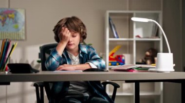 Çocukların odasında beyaz süveter ve mavi kareli gömlekli bir okul çocuğu ödevlerini yapıyor. Zor bir görev için acı çeken çocukları yazıyor.