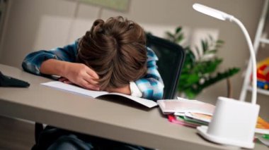 Çocuk odasında beyaz kazaklı ve mavi kareli tişörtlü bir çocuk başının arkasındaki masaya uzanmış. Sıkıntılı duygular çeken çocuklar için zor bir görev.