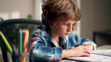 Beyaz kazaklı ve mavi kareli tişörtlü bir okul çocuğu çalışma odasında ödevlerini yapıyor ve acı çeken çocuklar için zor bir görev hazırlıyor.