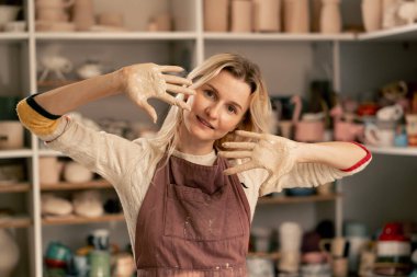 Kil modelleme stüdyosuna kapan kilden önlük giyen güzel kız kameraya gülümsüyor kirli kıyafetlere bakıyor hafta sonu hobileri kamerada.