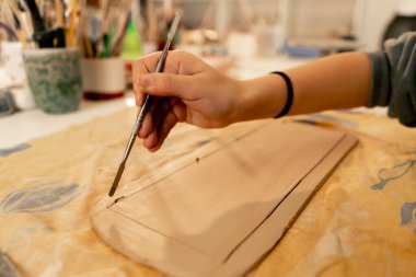 Bir mankenlik stüdyosuna yakın plan, kadın elleri kilden bir tabakta istenen şekli kesiyor, boş, cetvel ve tahta yığını.