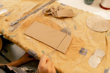 Bir mankenlik stüdyosuna yakın plan, kadın elleri kilden bir tabakta istenen şekli kesiyor, boş, cetvel ve tahta yığını.