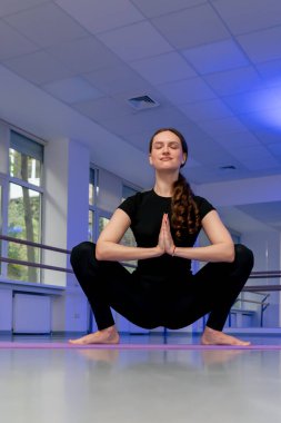 Yoga yüksek lisans sınıfının stüdyosunda mor hasırlı uzun saçlı kız elleri katlı bir kurbağa pozisyonunda oturuyor.