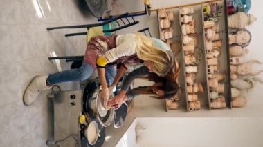 Kil modelleme stüdyosundaki dikey video çömlekçi çarkında kil şekillendiren genç kız elleri eski el sanatları öğretmenine yardım ediyor.