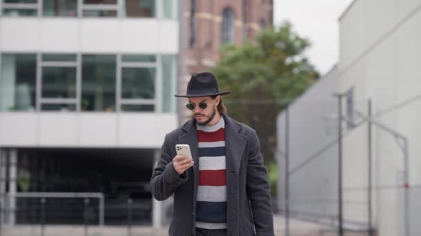 夏時間の屋外散歩中にスマートフォンを使用してコートやサングラスで幸せなハンサムなブルネットの男 アプリケーションによるタクシー注文を待っているスマート男 — ストック動画