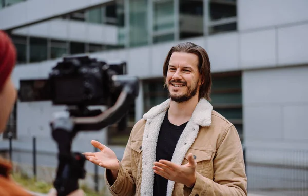 Long haired actor man talking gesturing while cameraman shooting him in urban street. Attractive male giving interview to the camera while working at the office building background