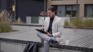 Takım elbiseli konsantre Avrupalı işadamı ofis binasının yanındaki bankta oturuyor ve dijital tablet kullanıyor. Uzun saçlı adam buluşmayı bekliyor, ayarlamalar yapıyor ve internette sohbet ediyor.