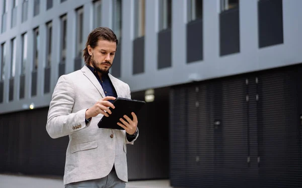 Professional employee writing notes on tablet while standing near the office building. Adult entrepreneur businessman creating new ideas and doing corporate tasks at his device. Business strategy