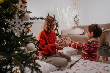 Evdeki güzel süslü Noel ağacının yanında otururken annesine Noel hediyesini sunan tatlı küçük çocuk. Hoş çocuk kış tatili boyunca annesiyle eğleniyor.