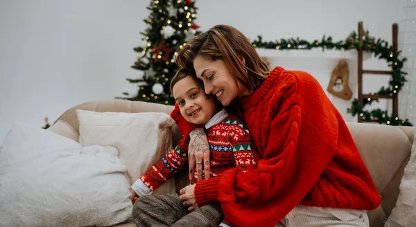 Noel ağacının yanında oğluyla kucaklaşan koyu renk saçlı genç bir anne Noel için dekore edilmiş bir odada. Aile ve Noel konsepti