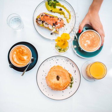 Top view of the unknown woman hand holding avocado toast. Person eating a sandwich with avocado, salmon, micro greens and drinking coffee on the breakfast in cafe