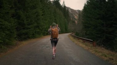 Back view of the young woman traveller walking in middle of empty road in mountains, carrying backpack, enjoying of the nature. Travel road trip wanderlust concept