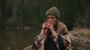 Tourist girl stopped near mountain river, drinking coffee against the backdrop of mountains. Woman warming up with hot beverage and enjoying of wild nature