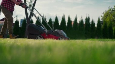 Cropped view of man mowing green grass in modern garden with lawn mower. Unknown man using lawn mower in the backyard. Professional lawn care service concept