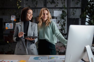 Women looking at the computer screen and discussing something at the office. Women colleagues standing with tablet at workplace 
