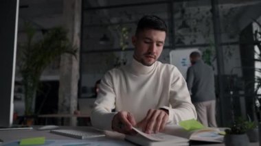Active businessman reading notes and searching information while working in the modern coworking office with colleagues at the background. Brunette office employee spending time at open space