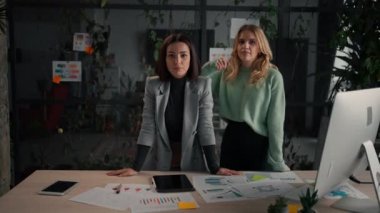 Two millennial women looking at camera confidently and posing in office during break. Young female colleagues standing at workplace
