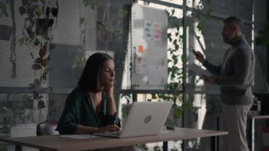 Attentive brunette businesswoman wearing green shirt looking laptop computer screen in open space coworking office. Her colleague working at the background