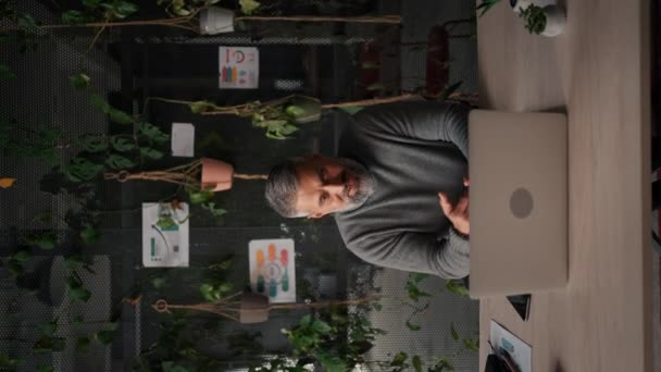 シニア男の垂直ビューは ビデオ通話中にノートパソコンの画面を振っています 現代の緑の植物オフィスのテーブルに座っている成熟したビジネスマン ウェブカメラを見て オンラインで話しています — ストック動画